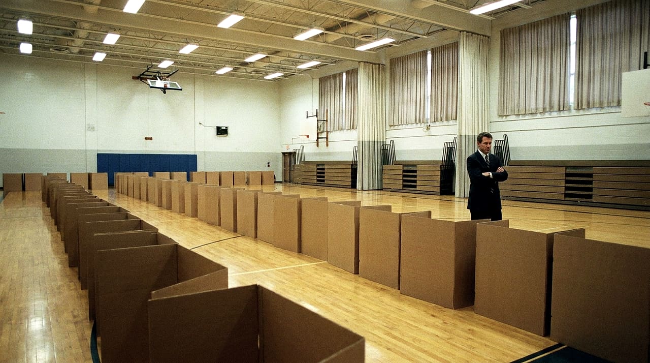 Rows of empty cardboard voting booths fill a school gymnasium as a man in a dark suit stands watching with his arms crossed.