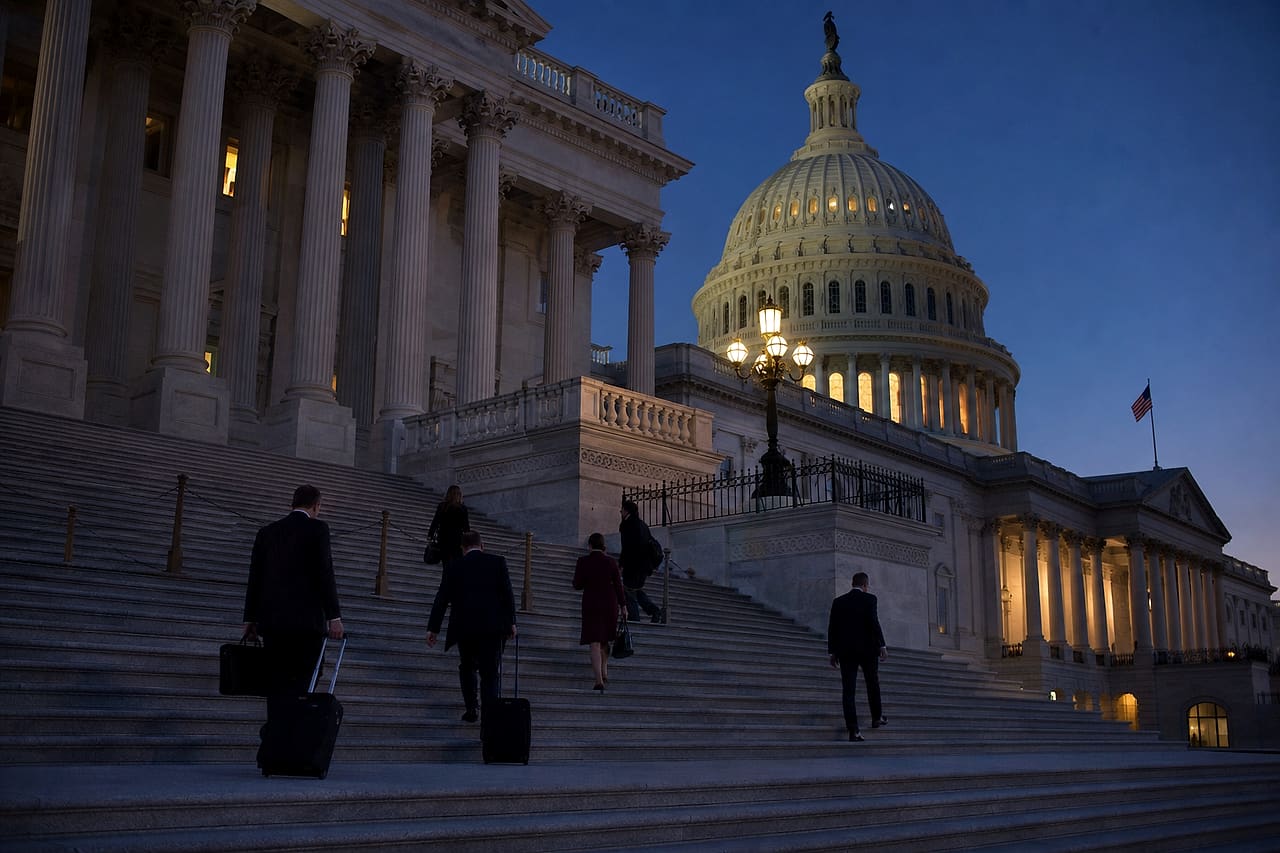 Five figures carry briefcases and rolling bags on the United States Capitol steps before sunrise.