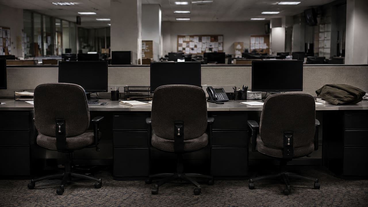 Three empty newsroom chairs face dark computer monitors beside a neatly folded unmarked military jacket.