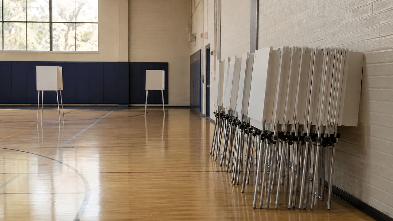 Folded voting booths are stacked beside the wall of an empty community-center gym, with a few booths still standing.