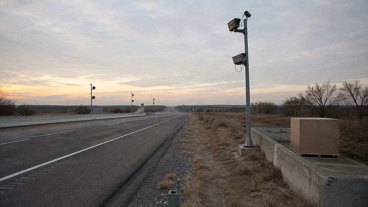 Anonymous surveillance cameras line an empty Texas highway beside a plain unopened shipping crate at dawn.
